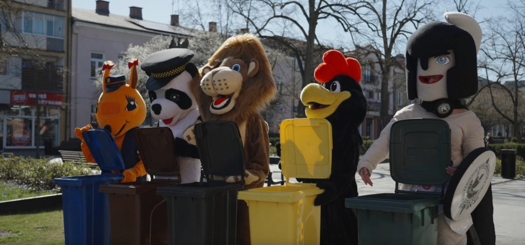 Pięć maskotek stojących przy kolorowych koszach na śmieci na deptaku, w przestrzeni publicznej